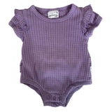 A baby's short-sleeve waffle-knit bodysuit in lavender purple, laid flat on a speckled stone countertop. The bodysuit features ruffled cap sleeves and a ribbed neckline.
