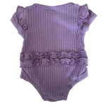 A lavender purple ribbed knit baby bodysuit with ruffled cap sleeves and a ruffled waistband, laid flat on a speckled white countertop.