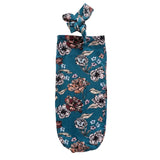 A dark teal fabric wine glass cover with a floral print, featuring a knotted top handle, laid flat on a speckled white surface.