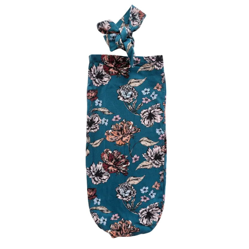 A dark teal fabric wine glass cover with a floral print, featuring a knotted top handle, laid flat on a speckled white surface.