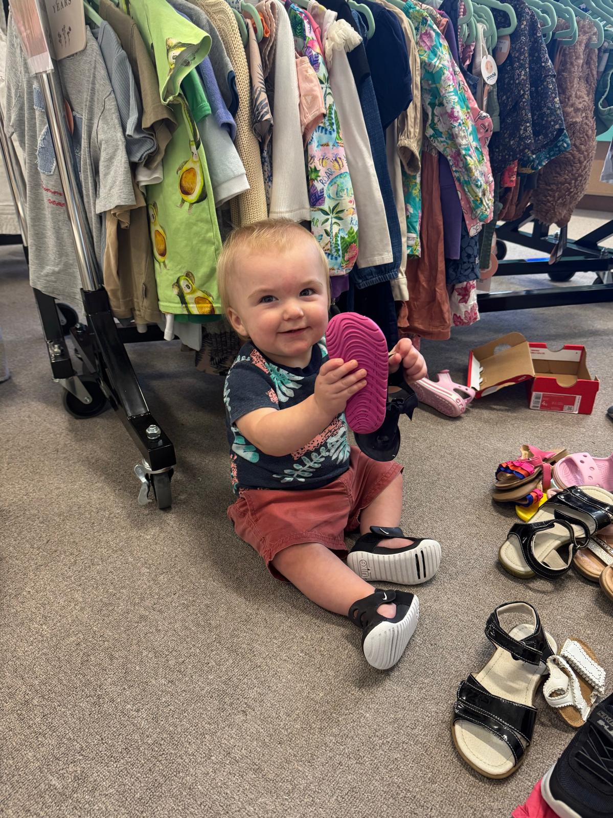 Baby sitting on the floor holding a pink shoe with clothes and shoes around